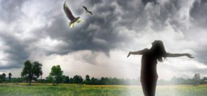A person with arms outstretched standing in a field under storm clouds with flying doves.