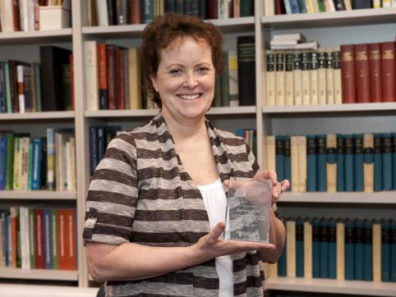 A smiling woman holding a glass award standing in front of a tall bookshelf filled with books.