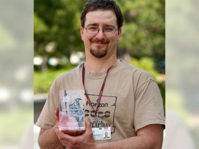 A smiling man holding a glass award with green trees in the background.