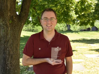 A smiling man in a red shirt holding a glass award, standing outside next to a tree.