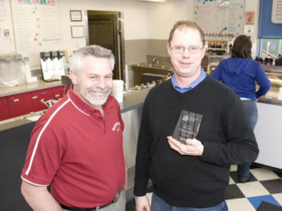 Two smiling men, one holding a glass award, standing in front of an ice cream counter.