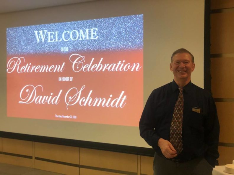 A smiling man standing in front of a screen with the text: Welcome to the Retirement Celebration in honor of David Schmidt.