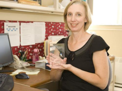 A smiling woman seated at a computer desk holding a glass award.