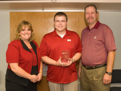 A smiling man in a red WSU shirt holding a glass award flanked by a smiling woman and man.