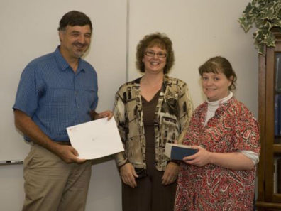 Three smiling people, one holding a blue box and another a certificate.