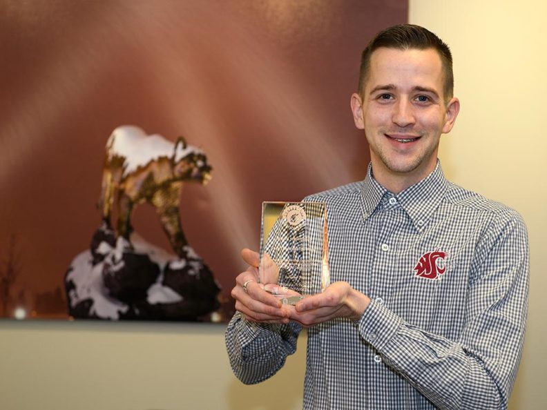 A smiling man holding a glass award with a large photo of a Cougar statue in the background.