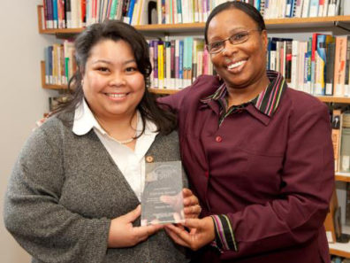 Two smiling women, one holding a glass award. A large bookcase in the background.