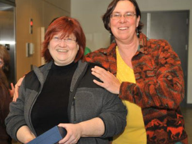 Two smiling women, one holding a blue box and the other with her arms on the other's shoulders.