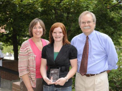 A smiling woman holding a glass award flanked by a smiling woman and man.