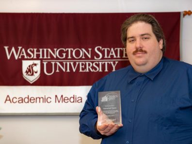 A smiling man holding a glass award in front of a red and white banner that reads: Washington State University Academic Media.