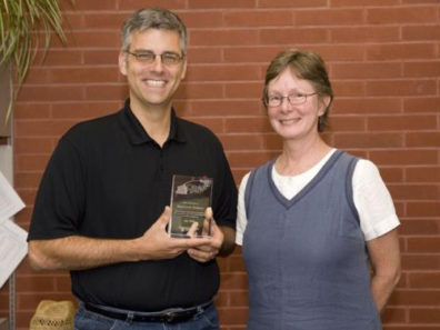 A smiling man and woman, the man is holding a glass award.