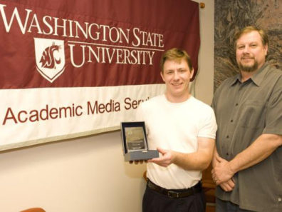 Two smiling men, one holding a glass award, standing next to a banner with the text: Washington State University Academic Media Service.