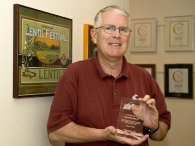 A smiling man holding a glass award next to a framed poster for the Lentil Festival.