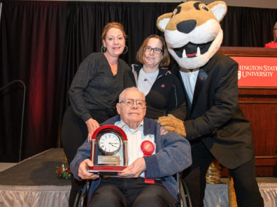 A seated man holding a clock award with two smiling women standing behind him next to a person wearing a WSU Cougar mascot costume and a suit.