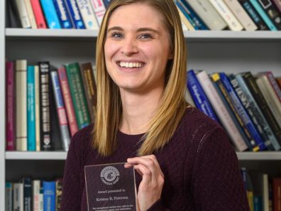 A smiling woman holding a glass award and standing in front of a bookshelf.