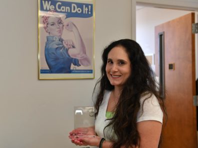 Smiling woman with long dark hair holding an award standing in front of a Rosie the Riveter poster.