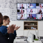 Photo of a meeting room with people applauding and other people on a video call.